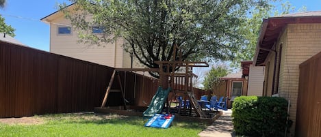 Fully enclosed back yard with a fire pit, grill, chairs, play set and corn hole.