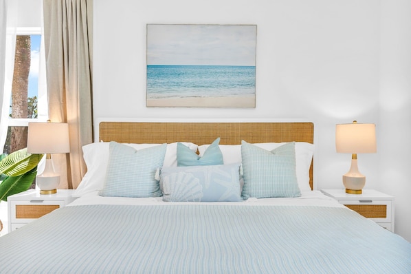 The primary bedroom suite is coastal "Serena & Lily" inspired. The king bed is a mango wood platform bed with a woven cane head and footboard in a natural finish, topped with a LUCID aloe gel memory foam mattress and 100% luxurious cotton linens.