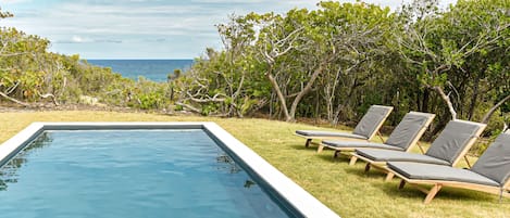 Vue sur l'océan et la piscine depuis le jardin d'une villa en bord de mer à Marie Galante, avec transats en bois confortables