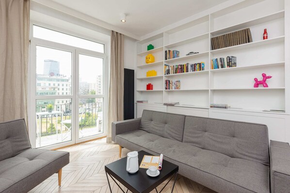 A bright living room with a grey sofa, modern shelves, and large windows offering a view of the city skyline.
