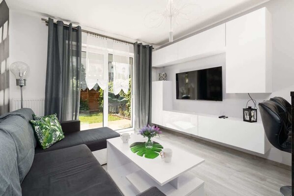 A modern living room with a large TV mounted on a white wall, a gray sofa, and a coffee table with fresh flowers.

