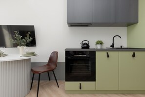 A compact kitchen with olive-green cabinets, black appliances, and a small round table with a chair.

