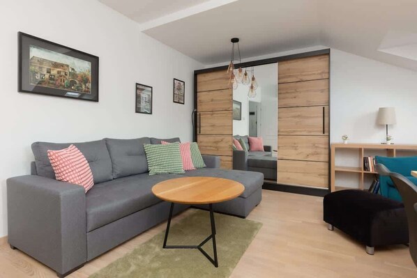A bright living area with a comfortable gray sofa, a round wooden coffee table, colorful pillows, and a partial view of a wooden closet.

