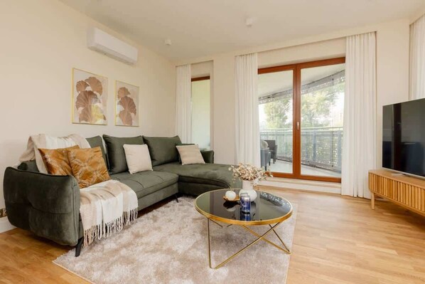 A spacious and bright living room with a comfortable green sectional sofa, a flat-screen TV, and large windows allowing natural light to flood the room.

