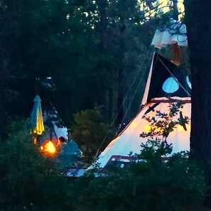 The Raven's Myth is a 16-foot Tipi on an oversized deck. It has outdoor chairs, a large umbrella, a propane fire pit: a picnic table, and a briquet BBQ grill.