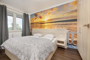 A spacious bedroom with a comfortable double bed, vibrant wall art, and large windows with curtains for privacy.

