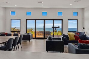 Spacious living area with large windows offering a scenic view of the ocean. The room features comfortable seating with colorful cushions, a dining table with chairs, and a telescope placed near the glass doors leading to an outdoor deck, suggesting a spa