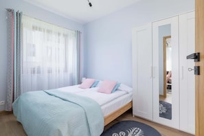 A serene bedroom with pastel bedding, mirrored wardrobe, and large window allowing plenty of natural light.