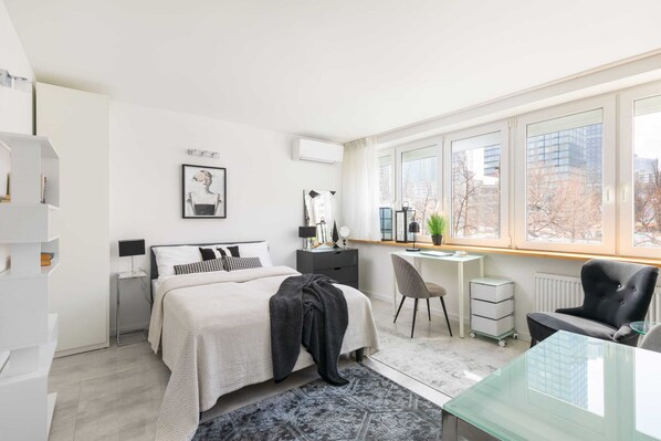 A bright and modern studio apartment with a comfortable bed, stylish furniture, and large windows offering plenty of natural light.

