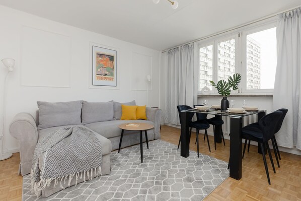 A bright living area with a gray sofa, a yellow accent pillow, and a small coffee table. Natural light pours in through patterned windows, creating a clean, modern atmosphere.
