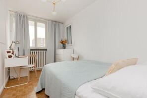 A white bedroom featuring a spacious bed, subtle beige accents, and a simple bedside table with a lamp. The overall style is minimal and light.
