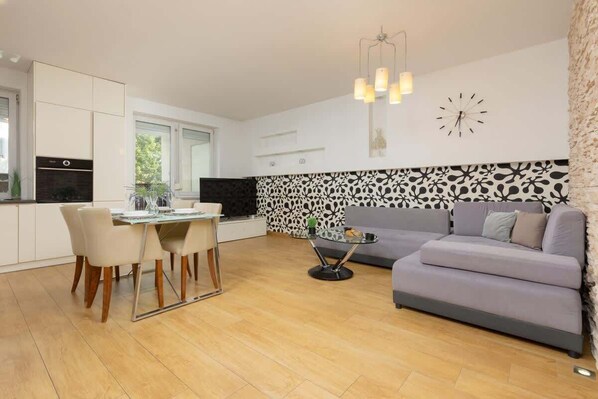 A spacious open-plan living and dining area featuring a gray sectional sofa, a wooden dining table with white chairs, and patterned wall decor for a modern and cozy ambiance.

