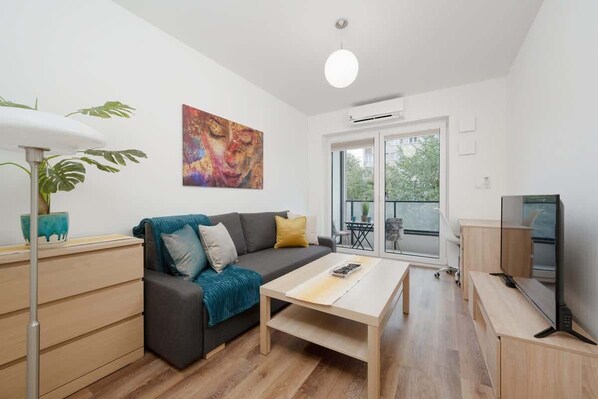 A bright living room with a gray sofa, yellow pillows, a wooden coffee table, and a large window letting in natural light.


