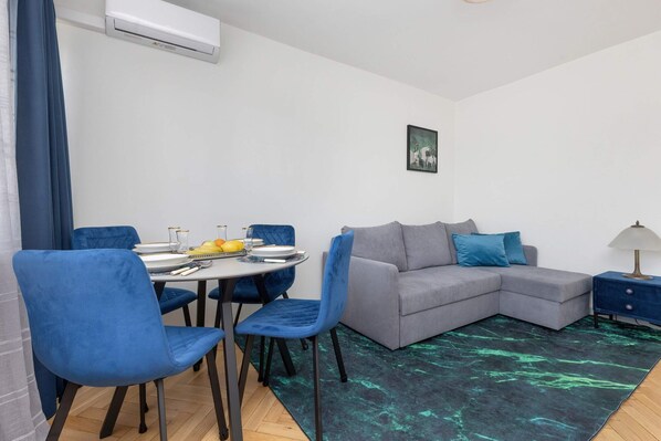 A living and dining area with a gray sofa, blue chairs, and a matching rug, complemented by natural light streaming through large windows.

