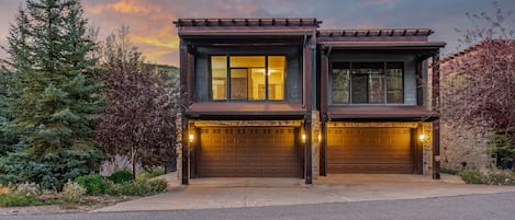 Modern mountain duplex with warm glowing interiors, rustic timber accents, and a peaceful setting framed by evergreens and twilight skies.