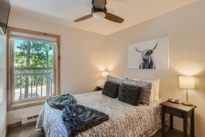 A cozy bedroom with a double bed, two bedside tables with lamps, a window with a scenic view, and a ceiling fan. The bed has a gray blanket and pillows, and a large artwork of an animal hangs above it.
