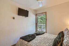 A small bedroom features a wall-mounted TV, a bed with patterned bedding and fur pillows, a ceiling fan, and a window with a view of trees and a balcony railing.